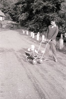 A man in a cable-knit sweater and trousers pushes a vintage rotary lawnmower along a rural, gravel road. Stone pillars line t...