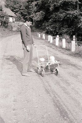 A young man in a sweater and trousers stands beside a vintage wicker baby carriage on a dirt road. Stone posts line the roads...