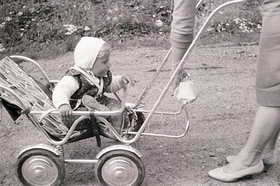 A young child, wearing a patterned dress and bonnet, sits in a vintage silver stroller, held by an adult. The stroller featur...