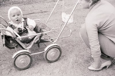 Black and white photograph shows a baby seated in a vintage, chrome-framed stroller. The baby wears a bonnet and formalwear. ...