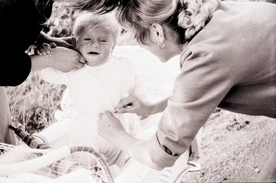 A distressed infant in a white dress is held by a woman with a distinctive hairstyle and hoop earrings. A stroller partially ...