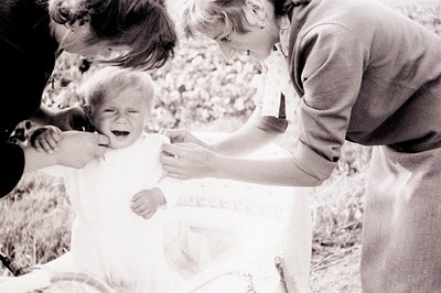 A crying baby in a white dress is being held by two adults. The scene appears to be an outdoor setting, likely a garden or pa...