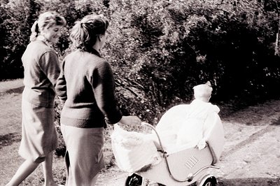 Two women stroll along a path, pushing a vintage pram with a doll inside. The scene appears to be captured in a garden or par...