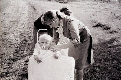 A young child sits in a modified cart or stroller, seemingly outdoors. The caregiver, dressed in a 1960s-style dress and patt...