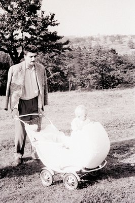 A man in a tweed suit pushes a vintage carriage containing a baby on a grassy field, backed by a gentle hillside. The carriag...