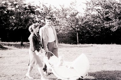 A young couple strolls through a grassy area, pushing a vintage wicker baby carriage. The woman wears a tailored suit, and th...