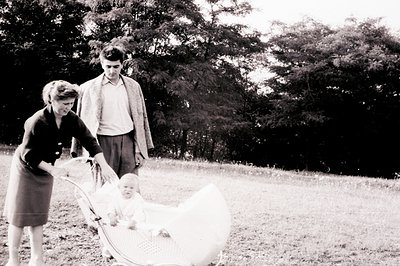 A couple strolls through a grassy area, pushing a wicker baby carriage. The woman wears a modest dress and the man a tweed ja...