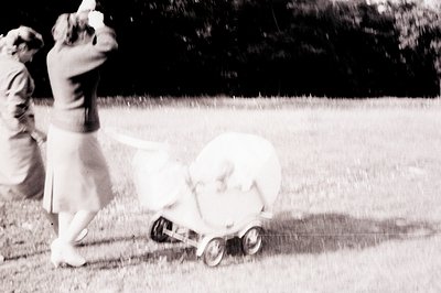 Vintage black and white shot captures a woman in a 1960s-style dress, pushing a vintage baby carriage across a grassy lawn. D...