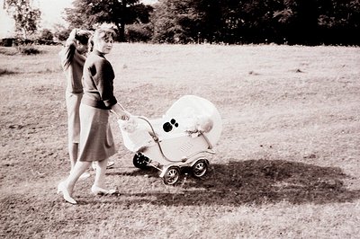 A woman in a 1960s-style dress and heels pushes a vintage, ornate baby carriage across a grassy field. Another figure is part...