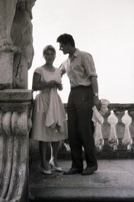 A stylish couple poses near an ornate stone balustrade. The woman wears a patterned dress & heels; the man, a short-sleeved s...
