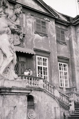 Elegant, aging estate facade with a sculpted figure overlooking tiered steps and a balcony where two figures stand. Building ...