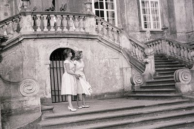 Two young women in matching knee-length dresses and heels pose on a grand stone staircase. The ornate architecture features a...