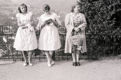 Three women stand by a wrought-iron railing overlooking a landscape. All wear patterned summer dresses and carry accessories:...