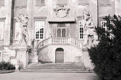 Formal, symmetrical architecture features a central entrance flanked by dramatic allegorical statues. Stone steps lead to an ...