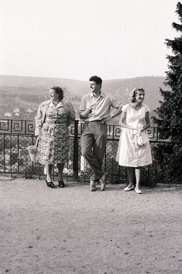Three figures pose outdoors along a railing with a hillside view. The woman on the left carries a wicker basket. The man wear...