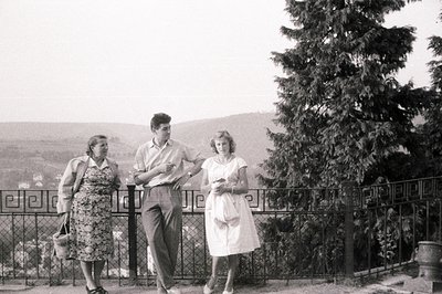 A relaxed group portrait: A woman in a dress and jacket, a young man in slacks & short-sleeved shirt, and a woman in a floral...