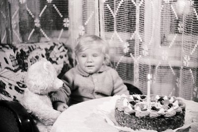 A young child sits next to a large teddy bear and a birthday cake with lit candles. The scene is framed by patterned curtains...