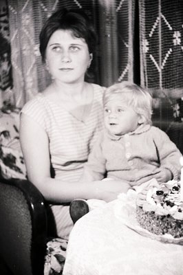 A seated woman holds a young child, both appearing to gaze directly at the camera. A decorated cake with lit candles sits on ...