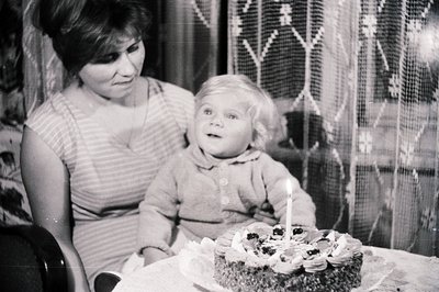 A young girl sits before a decorated cake with a single candle, observed by a woman with a striped dress. Interior scene, lik...