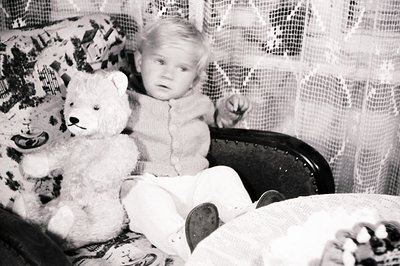 Young child seated in an ornate chair, clutching a worn teddy bear. Dressed in a knitted sweater and trousers, posed formally...