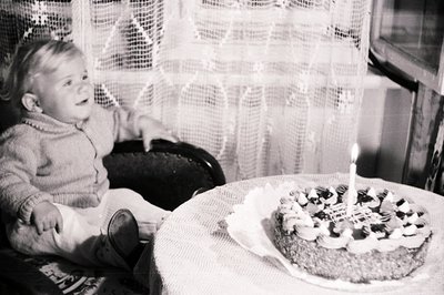 A young child sits in a dark wicker chair, gazing at a layered cake adorned with cream frosting and a single lit candle. The ...