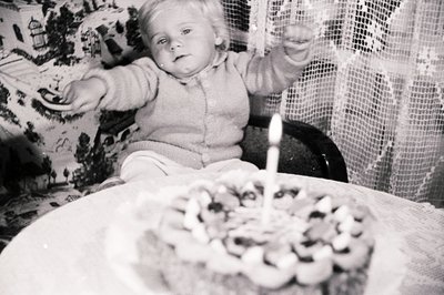 A young child sits in a highchair, reaching for a frosted cake with a single lit candle. The setting appears to be a domestic...