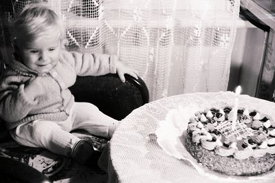 A young child, likely a toddler, sits in a cushioned chair, observing a decorated cake with a single lit candle. The scene su...