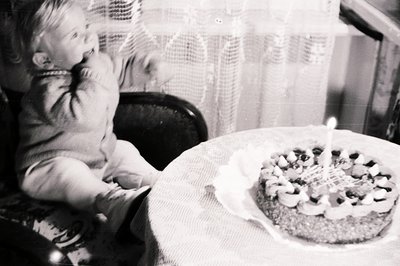A young child, likely a toddler, sits in a high chair, reaching towards a decorated cake with a single lit candle. The cake s...