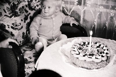 A young child, possibly a toddler, sits in an ornate armchair, appearing to observe a cake decorated with what appear to be s...