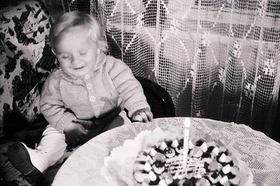 A young child, seated at a table covered with a lace tablecloth, gazes towards a small cake adorned with a single lit candle....