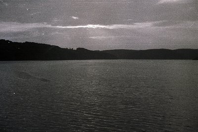 Moody black and white landscape with a still body of water reflecting an overcast sky. A dark shoreline and dense trees defin...