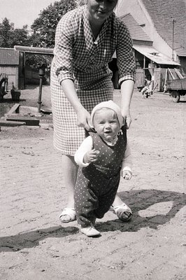 A young child in a polka-dotted overalls and bonnet takes tentative steps while being supported by an adult's hand. Farmhouse...