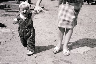 A toddler in a patterned romper and bonnet takes tentative steps, held by an adult's hand. Visible are the adult's floral pri...