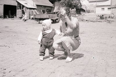 A young child in overalls and cap takes tentative steps outdoors. A woman in a checked dress squats, blowing bubbles. Rural s...