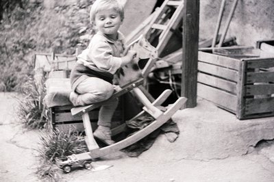 A young boy sits on a wooden rocking horse, smiling. He wears overalls and a collared shirt, suggesting a mid-20th century, l...