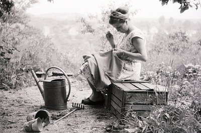 A woman seated on a wooden crate, focused on mending fabric with a needle. Gardening tools, including a watering can and smal...
