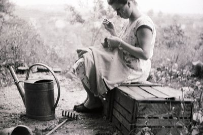 A woman sits outdoors, engaged in needlework on a wooden crate. A watering can and gardening tools rest nearby, suggesting a ...
