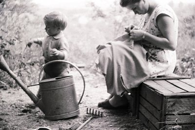 A young boy stands near a watering can, while an older woman sits on a wooden crate, seemingly engaged in needlework. Rustic ...