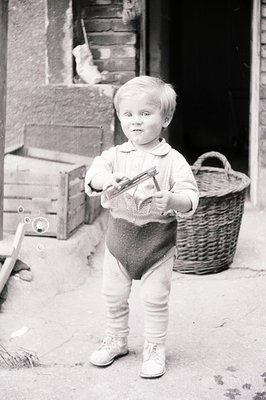 A young boy, standing outdoors, holds a small, antique-style camera. He's dressed in a collared shirt, bloomers, and shoes. A...