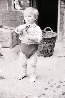 A young boy stands outside a building, wearing knit stockings, shorts, and a collared shirt. He poses with a playful expressi...
