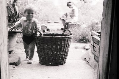 A young child in short overalls stands beside a large, woven basket, seemingly offering it. A woman, wearing a light-colored ...