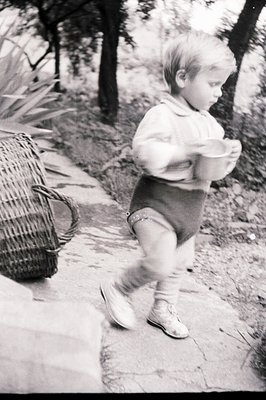A toddler walks along a cracked stone path, carrying a small metal pail. Subject wears short pants, a long-sleeved shirt & sn...