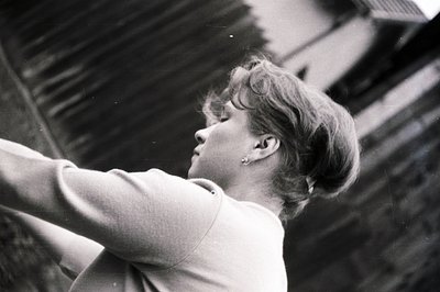 A young woman with short, styled hair is captured in profile, arms raised, against a weathered wooden building facade. The im...