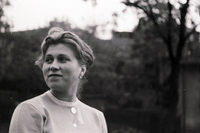 Portrait of a woman with a gently smiling expression, styled hair, and a simple, buttoned blouse. Captured outdoors, with blu...