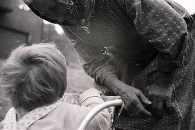 A black and white photo shows a young child, viewed from behind, seated in a high chair. An adult, likely a caregiver, is adm...