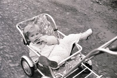 A young child, likely a toddler, sits in a vintage chrome and fabric stroller. The child wears a patterned dress and boots, g...