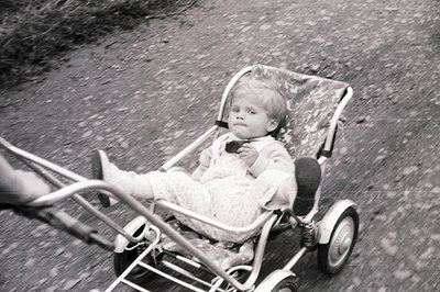 A young child sits in a vintage, metallic-framed wagon or stroller, captured in motion. The child wears a patterned knit outf...