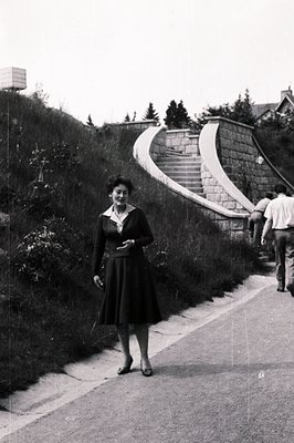 A well-dressed woman stands on a roadside alongside a large stone staircase leading up a hillside. The architecture suggests ...