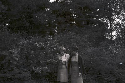 Two women in knee-length dresses and jackets stroll through foliage, partially obscured by dense trees. Visible fashion detai...