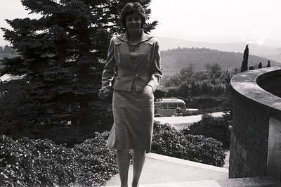 Elegant woman in a tailored suit descends stone steps, overlooking a sprawling landscape with a vintage bus visible below. Li...